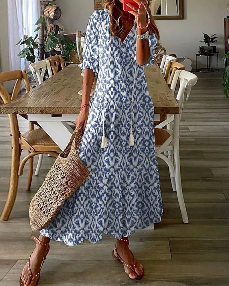 Woman in blue and white ikat maxi dress holding a woven straw bag, standing in a modern dining room with wooden table and chairs. Fashion, summer style.