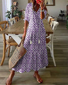 Woman in a purple ikat print maxi dress holding a woven straw bag, standing in a stylish dining room with wooden table and chairs. Fashionable summer outfit.