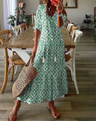 Woman in green patterned maxi dress holding a woven straw bag, standing in a stylish dining room with wooden table and chairs, casual summer fashion.