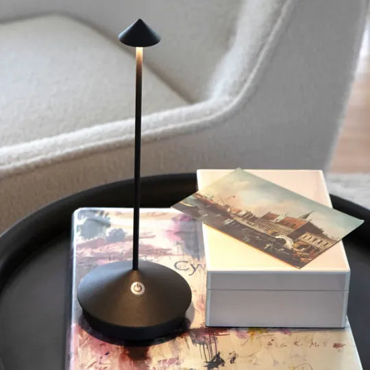 Modern black LED desk lamp on a round table, next to a white box and a postcard, in a cozy living room setting. Ideal for home office lighting.