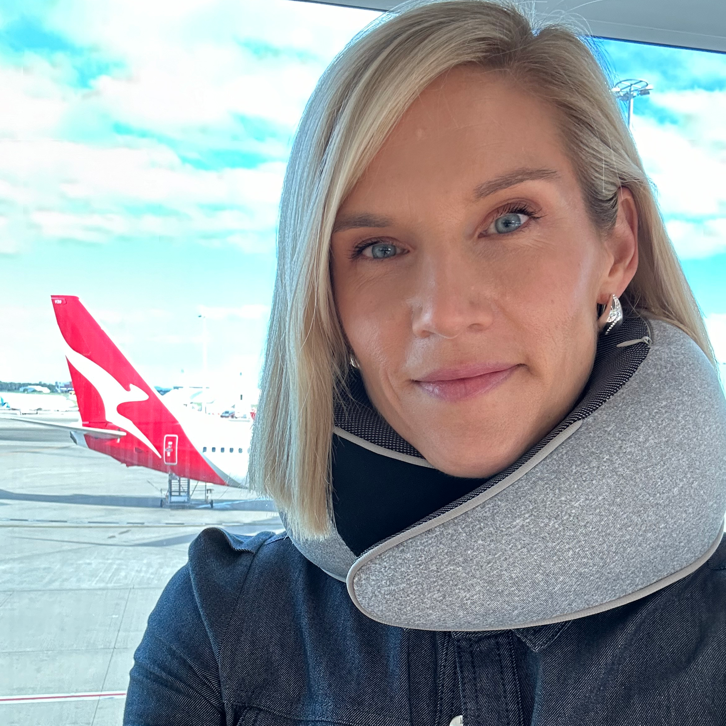 A person wearing a gray travel neck pillow at an airport with a Qantas airplane in the background, ideal for travel comfort and support.