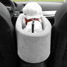 Small white dog in a gray knitted car seat booster, wearing a red harness, sitting between black car seats. Pet travel accessory, dog safety.