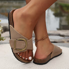 Beige slide sandals with large gold buckle, cork sole, and white pedicure, worn by a person with an anklet. Fashionable summer footwear.