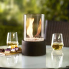 Tabletop fire pit with glass cylinder, surrounded by wine glasses and snacks on a patio table. Perfect for outdoor ambiance and garden decor.