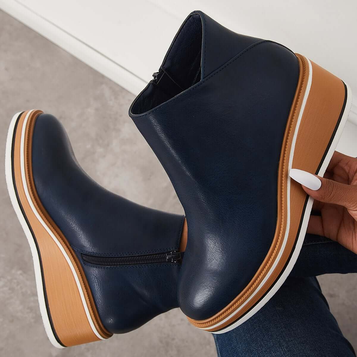 Navy blue wedge ankle boots with tan and white platform sole, side zipper detail, held by a hand with white nails on a neutral background.