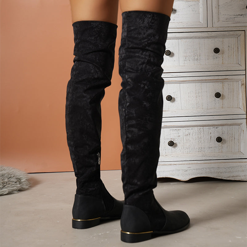 Black over-the-knee suede boots with low heels, worn by a person standing on a beige floor, next to a white wooden dresser. Fashion footwear.