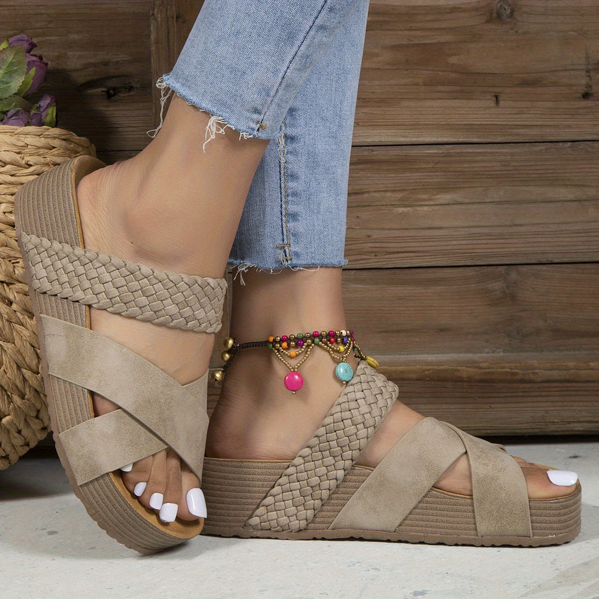 Beige woven platform sandals with crisscross straps on a woman's feet, paired with light blue jeans and a colorful anklet. Fashion footwear.