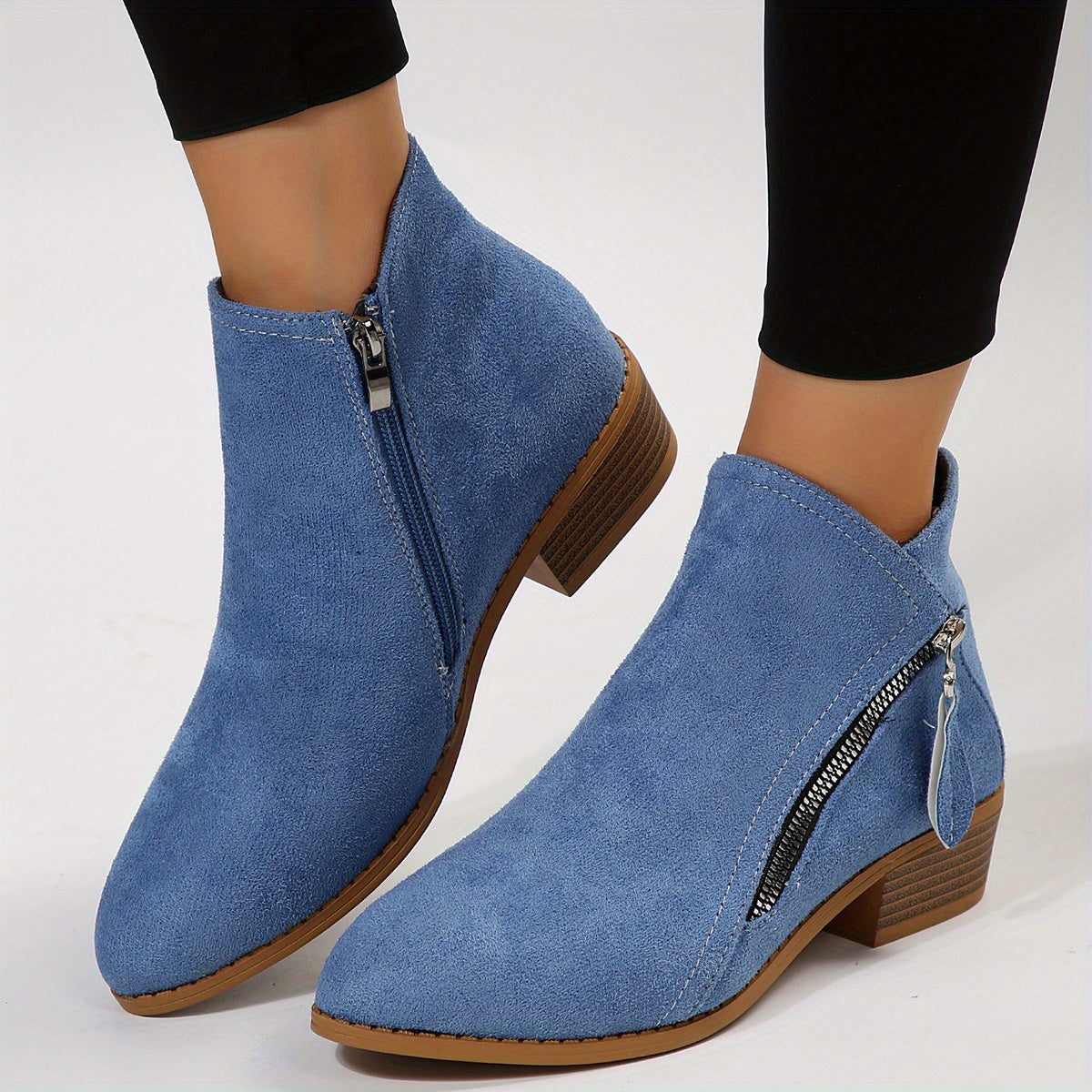 Blue suede ankle boots with side zippers, featuring a low wooden heel, worn with black leggings. Fashionable women's footwear for casual style.