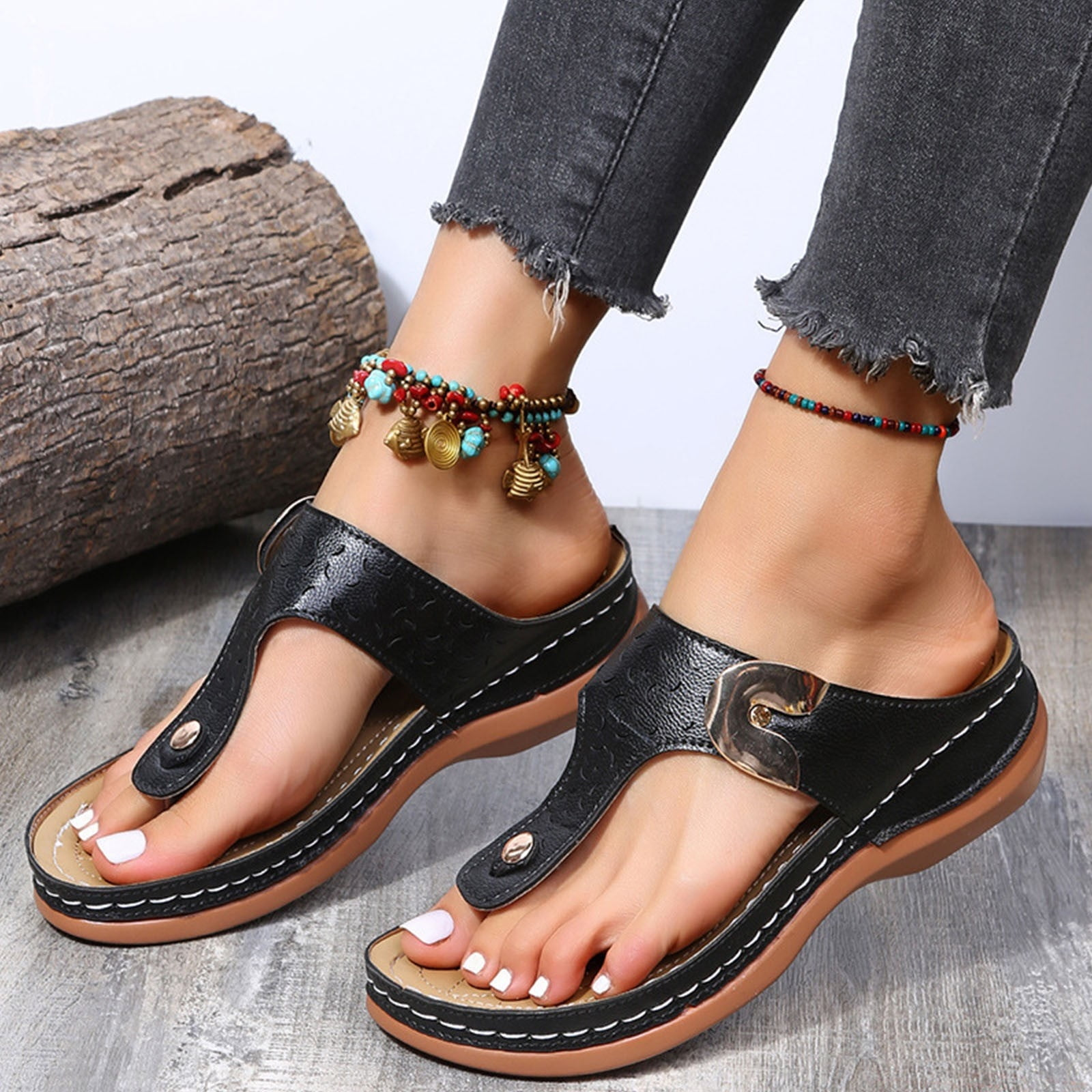 Women's black leather sandals with buckle, worn with beaded anklets and distressed jeans, on a wooden floor. Fashionable summer footwear.