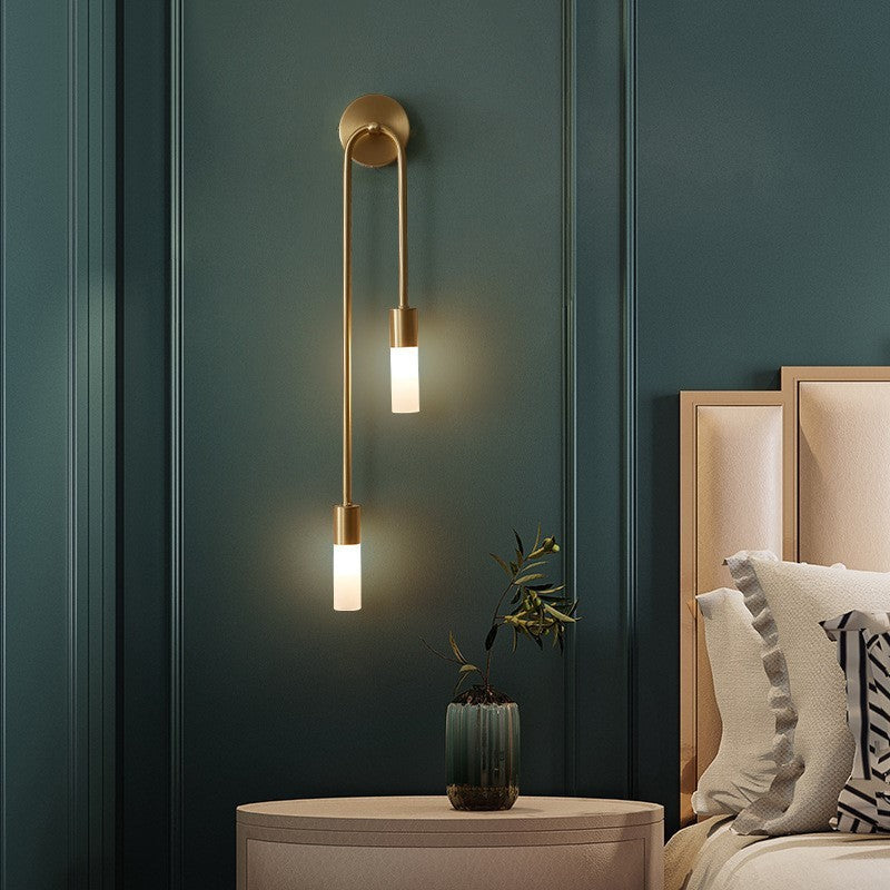 Modern gold wall sconce with dual frosted glass lights on teal wall, above round bedside table with plant, next to beige upholstered headboard.