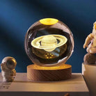 3D Saturn crystal ball lamp on wooden base, glowing in dark room, surrounded by astronaut figurines. Perfect space-themed decor gift.