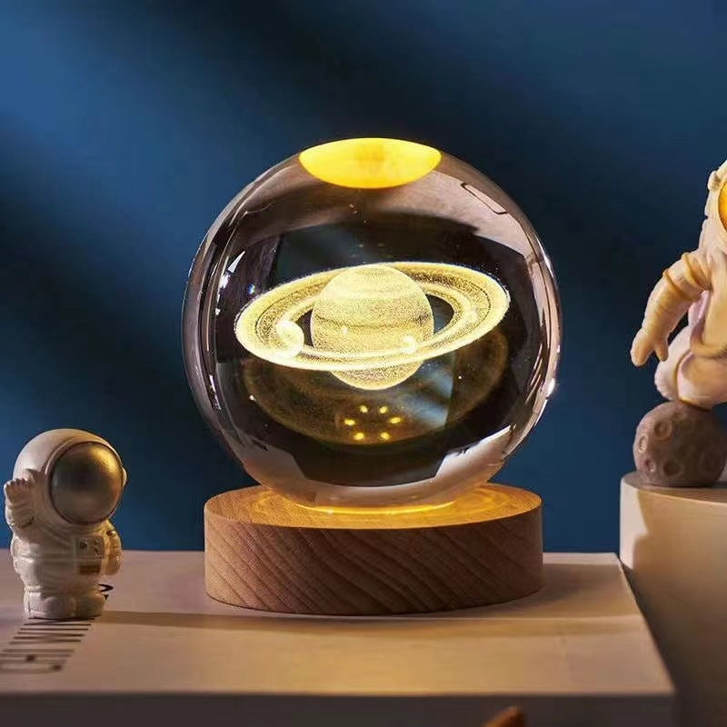 3D Saturn crystal ball lamp on wooden base, glowing in dark room, surrounded by astronaut figurines. Perfect space-themed decor gift.