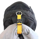 Black backpack with multiple zippers, featuring a yellow strap and buckle attachment holding a gray fabric. Durable travel backpack accessory.