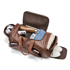 Brown leather duffel bag with open compartments showing packed items: headphones, camera, water bottle, clothes, shoes. Ideal for travel and gym use.