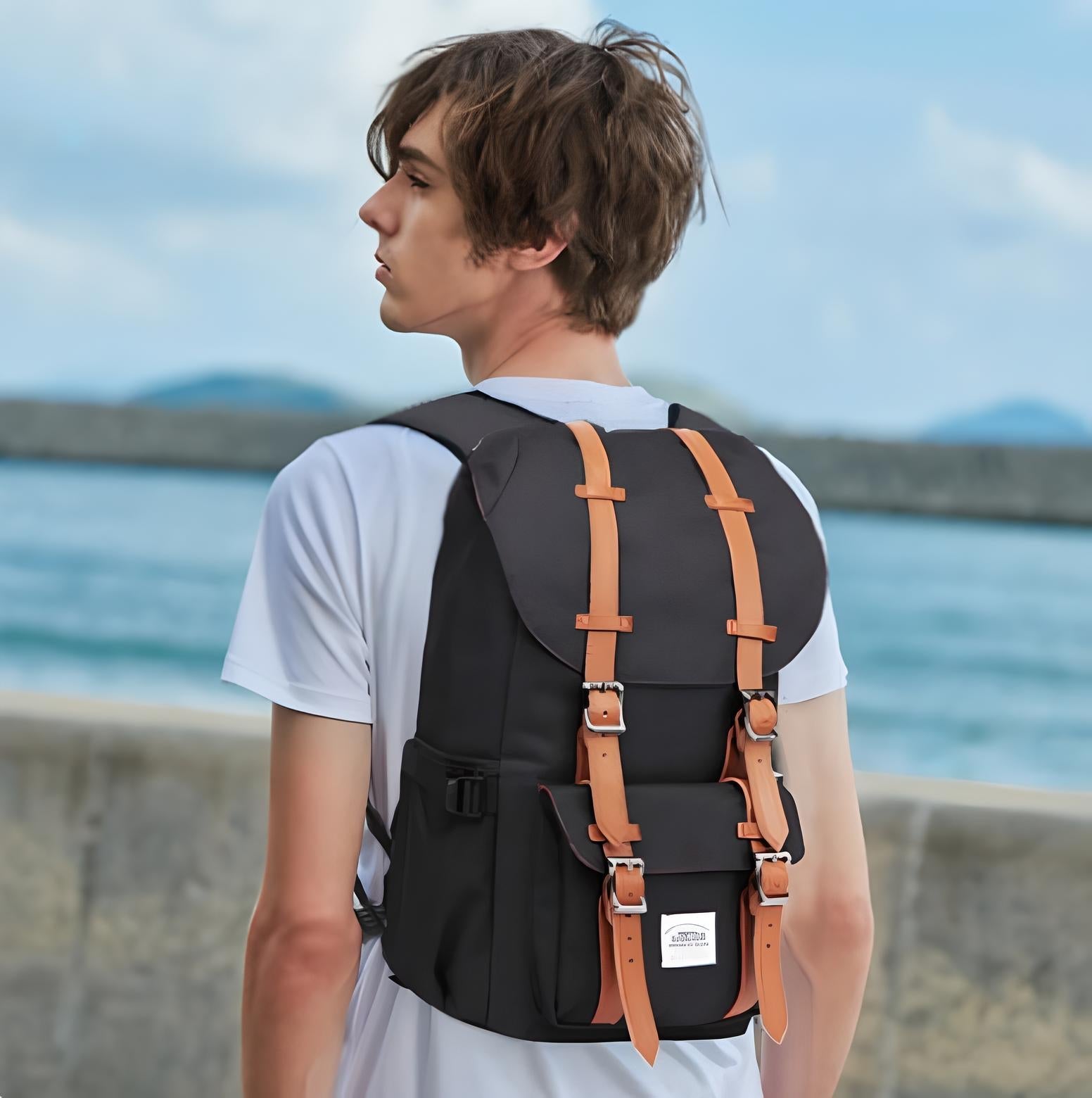 Man wearing a stylish black backpack with brown leather straps, standing outdoors by the sea. Trendy travel gear, perfect for hiking and urban adventures.