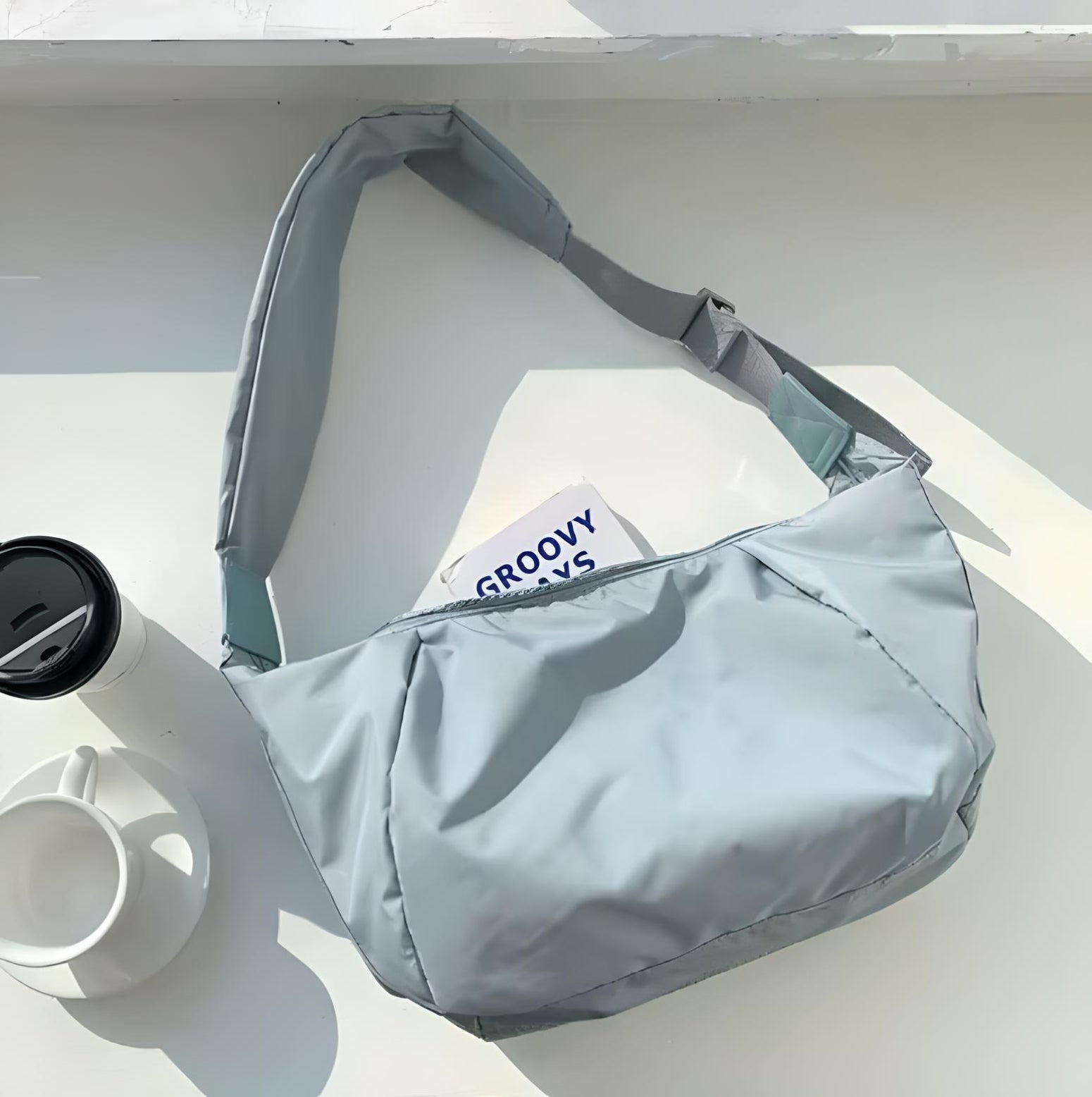 Light gray shoulder bag on white surface with adjustable strap, next to a black coffee cup lid and white mug. Stylish, modern accessory.