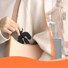 Woman placing portable handheld fan into beige handbag, with suitcase in background. Travel essentials, compact cooling device, stylish accessory.