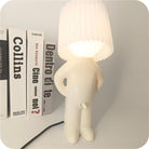 Quirky white lamp with a pleated shade shaped like a person, standing beside books on a shelf. Unique home decor, modern lighting design.