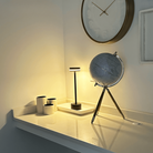 Modern desk setup with a sleek black lamp, decorative globe on a tripod stand, and minimalist white cups on a white surface, under a wall clock.