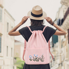 Woman wearing a straw hat and black dress with a pink drawstring backpack featuring floral embroidery, walking in an urban setting.