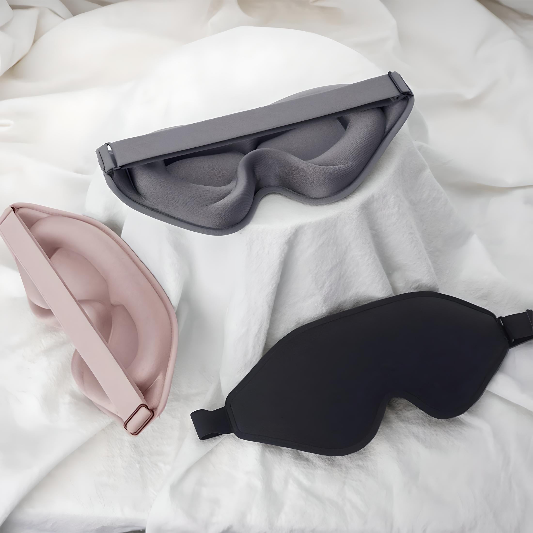 Three ergonomic sleep masks in black, gray, and pink on a white fabric background, designed for comfort and light blocking. Perfect for restful sleep.