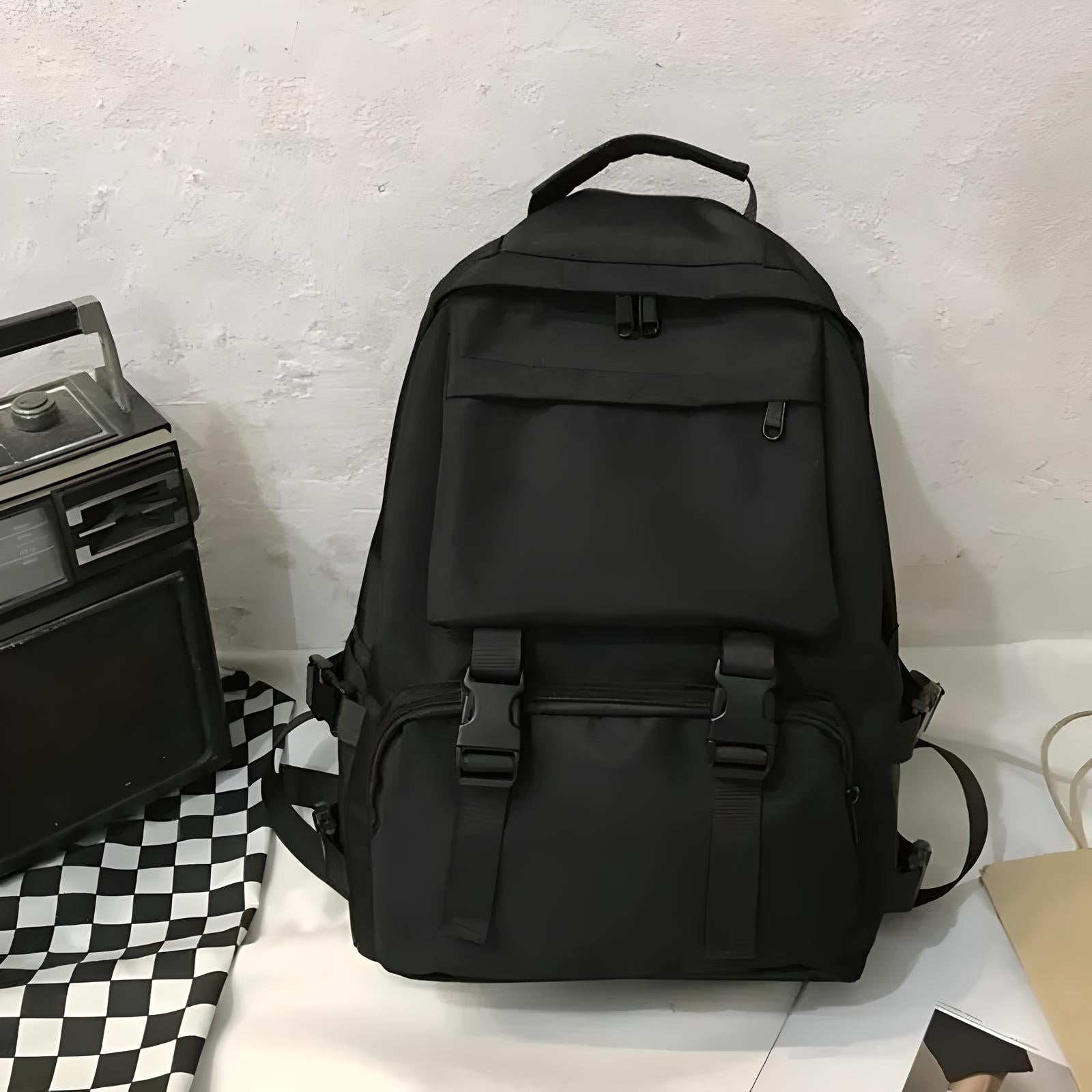 Black backpack with multiple compartments and buckles, placed on a checkered cloth beside a vintage radio. Ideal for travel and daily use.