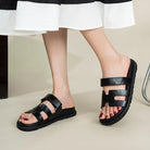 Black leather slide sandals with double straps on woman's feet, featuring a comfortable flat sole. Perfect for casual summer fashion and footwear trends.