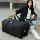 Woman sitting with large black wheeled duffel bag, wearing casual outfit. Travel luggage, durable design, convenient for trips.