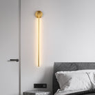 Modern gold wall sconce with vertical LED light, mounted beside a bed in a minimalist bedroom. Elegant lighting fixture for contemporary interiors.