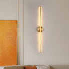 Modern vertical wall sconce with warm LED lighting, gold finish, and minimalist design, mounted on a beige wall beside an abstract orange painting.