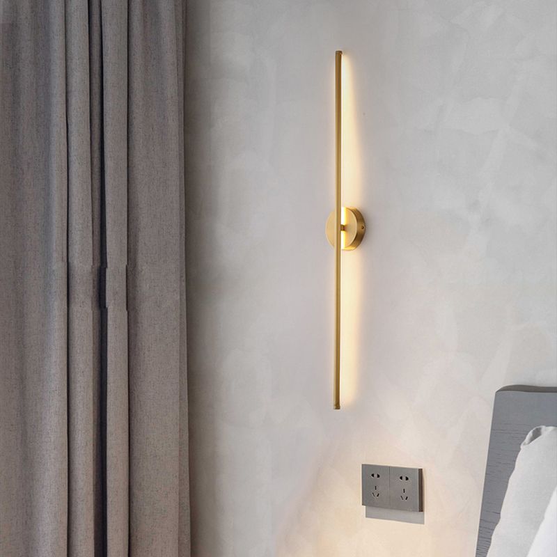 Modern gold wall sconce with vertical LED light, mounted on textured gray wall beside curtains and power outlet, minimalist bedroom decor.