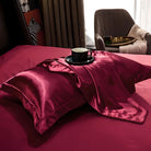 Luxurious red silk bedding set with two pillows and a matching throw, featuring a black coffee cup on a saucer, elegant bedroom decor.