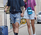 Couple walking with luggage, wearing casual summer outfits and crossbody bags, in an urban setting. Travel, fashion, street style, summer vacation.