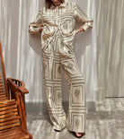 Woman wearing a beige cactus print pajama set, standing in a sunlit room with wooden chair. Stylish loungewear, comfortable sleepwear fashion.