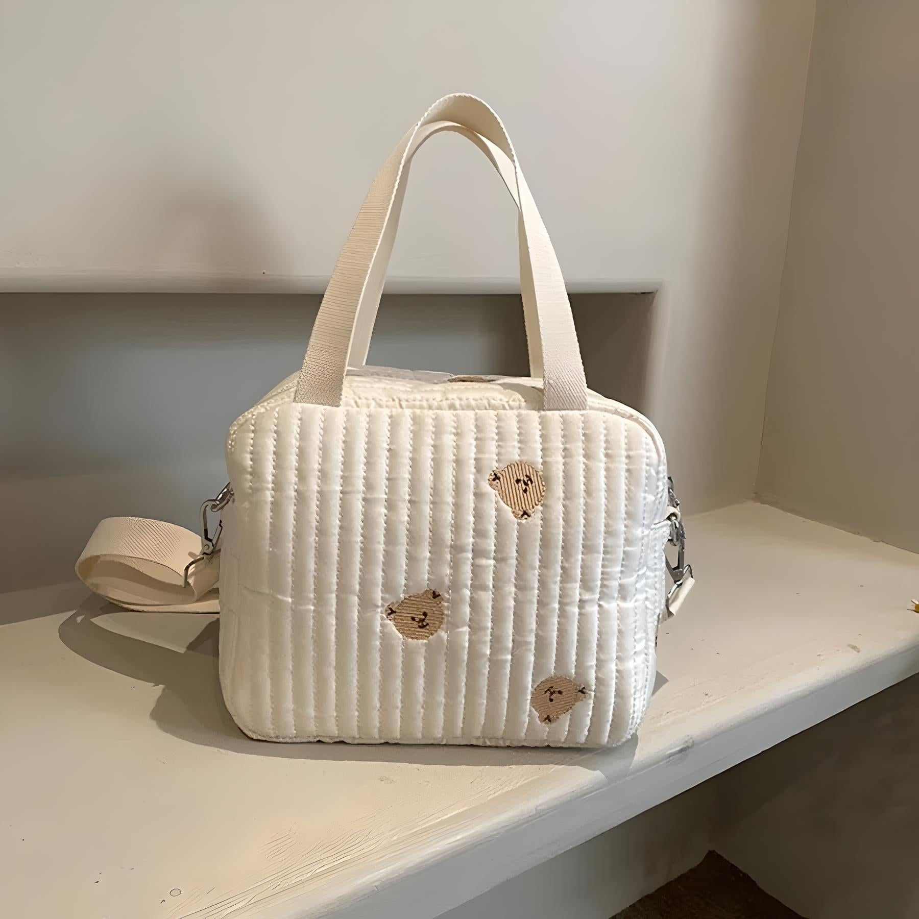 Quilted white handbag with beige handles and subtle embroidered heart designs, resting on a shelf. Fashionable, stylish accessory for women.