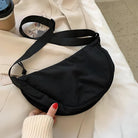 Black crescent shoulder bag with adjustable strap on a beige coat, next to a coffee cup and skincare product; stylish accessory for casual outfits.