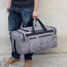 Man holding a stylish gray duffel bag with black straps, featuring multiple zippered compartments, ideal for travel and gym use. Casual jeans and shoes.