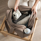 Person packing a beige travel bag with a camera, clothes, and a water bottle on a wooden stool. Travel essentials, packing, organization.