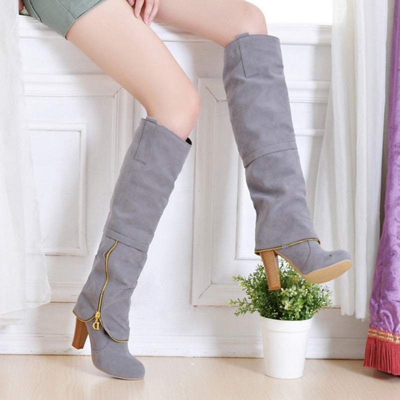 Gray knee-high suede boots with wooden block heels and gold zipper detail, worn by a seated person, next to a small potted plant.