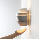 Modern white wall sconce with layered design, illuminating a hand beneath. Contemporary lighting fixture, minimalist home decor, soft ambient light.