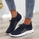 Black knit slip-on sneakers with white soles, featuring breathable mesh design and lace-up detail, worn with distressed denim jeans on wooden floor.