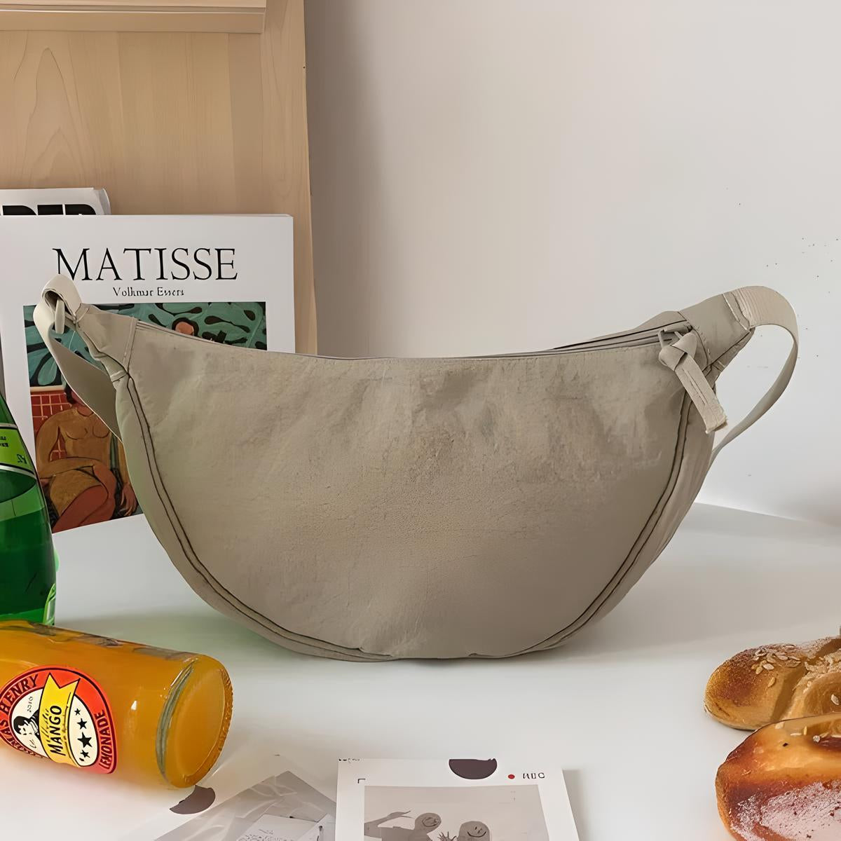 Beige crescent shoulder bag on a table with Matisse book, orange soda, and bread. Stylish minimalist accessory for casual fashion.