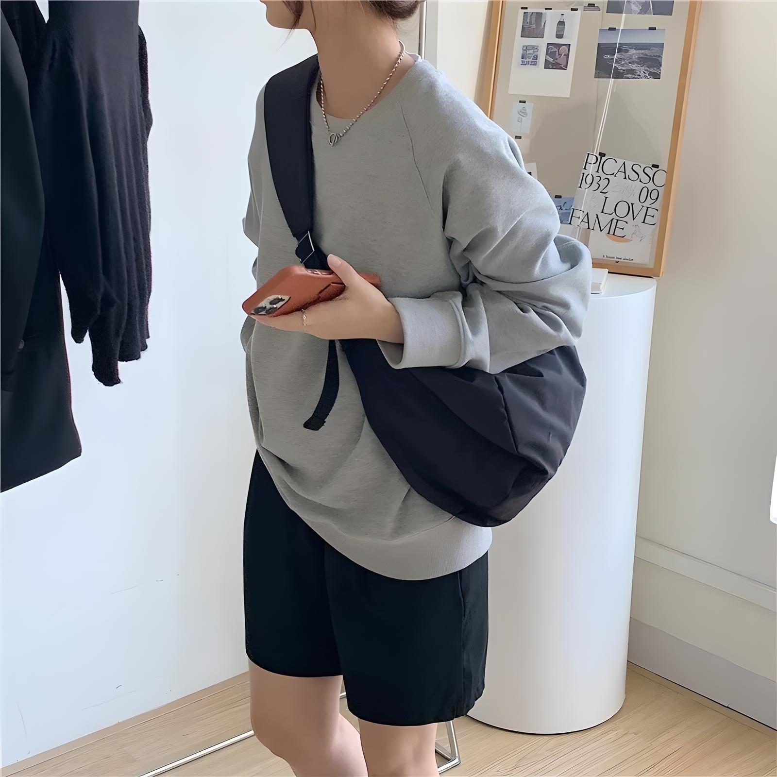 Woman in casual gray sweatshirt and black shorts holding a smartphone, wearing a black crossbody bag. Modern minimalist fashion, indoor setting.