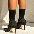 Black satin sock boots with high stiletto heels on a white textured background, showcasing elegant footwear fashion for women.