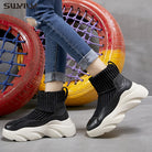 Black knit sock sneakers with chunky white soles, worn with rolled-up jeans, in front of colorful tire swings. Fashionable footwear, casual style.