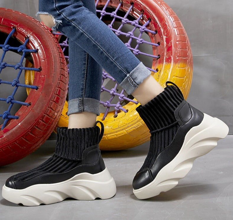 Black knit sock sneakers with chunky white soles, worn with blue jeans. Trendy footwear against colorful tire backdrop. Fashionable casual shoes.