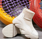 White high-top sneaker with ribbed sock design, chunky sole, and modern aesthetic, displayed against colorful tire backdrop. Fashion footwear.