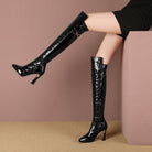 Black patent leather knee-high boots with stiletto heels, featuring a crocodile texture and gold chain detail, worn by a model in a black dress.