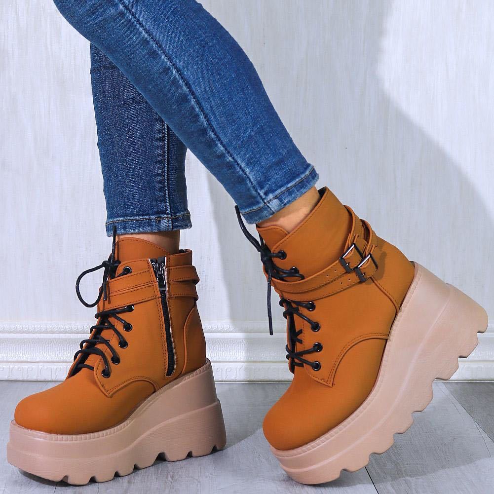 Women's tan platform ankle boots with chunky soles, lace-up front, side zipper, and buckle detail, paired with blue skinny jeans on a wooden floor.