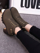 Olive green lace-up ankle boots with chunky heels, gold eyelets, and buckle detail, worn with black pants on a gray carpet. Fashion footwear.